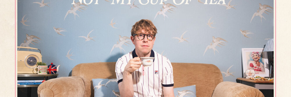 Josh Widdicombe holding a tea cup, sitting on beige satin sofa behind a coffee table with a china tea set on.